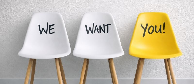 Drei Stühle vor einer weißen Wand mit der Aufschrift „We want you“ als Symbol für offene Stellen und Karrierechancen.